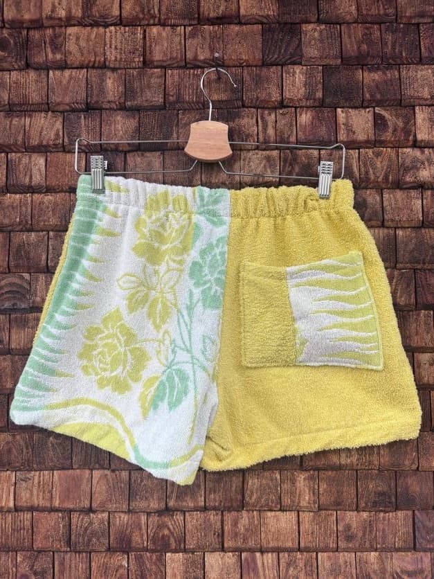 Rückansicht der Upcycling-Frotteeshorts „Yellow Rose ‘100“ von Old Wave auf einem Kleiderbügel vor einer Holzwand. Die Rückseite präsentiert das florale Rosenmuster in Weiß-Gelb-Grün auf der linken Gesäßhälfte, während die rechte Gesäßhälfte komplett in Gelb gehalten ist und eine eingenähte Tasche mit gestreiftem Retro-Muster besitzt.