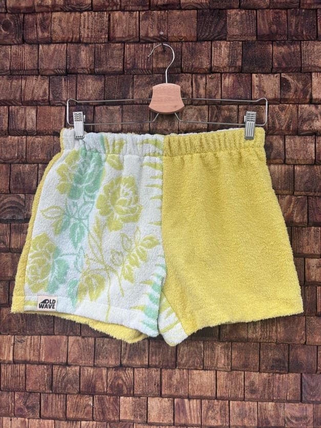 Vorderansicht der Upcycling-Frotteeshorts „Yellow Rose ‘100“ von Old Wave auf einem Kleiderbügel vor einer rustikalen Holzwand. Das fröhliche Design kombiniert ein linkes Hosenbein aus hellweißem Vintage-Frottee, das mit großen, blassgelben und mintgrünen Rosenmotiven verziert ist und das Old Wave-Logo am Saum trägt, mit einer unifarbenen, leuchtend gelben rechten Hosenhälfte.