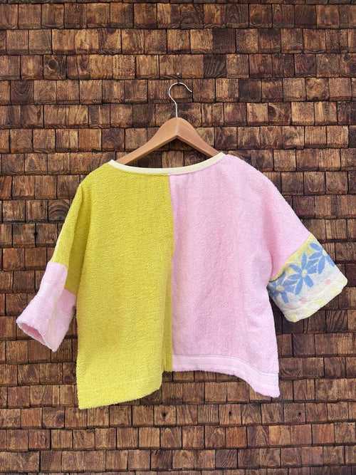 Rückansicht des Upcycling-Frotteetops „Sunfade Blossom Top ‘93“ von Old Wave auf einem Kleiderbügel vor einer Holzwand. Die Rückseite präsentiert ein markantes, hälftig geteiltes Color-Blocking-Design: Die linke Rückenhälfte ist in hellem Gelb mit einem rosa Ärmel gestaltet, während die rechte Rückenhälfte in Pastellrosa gehalten ist und einen Ärmel mit dem blaugrauen Retro-Muster besitzt.
