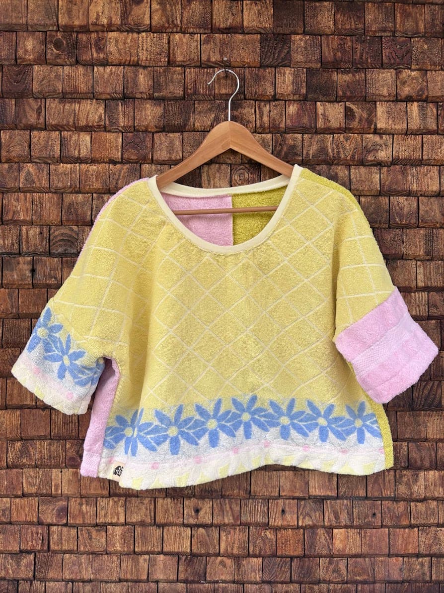Vorderansicht des Upcycling-Frotteetops „Sunfade Blossom Top ‘93“ von Old Wave auf einem Kleiderbügel vor einer rustikalen Holzwand. Der Rumpf des locker sitzenden Oberteils ist vorwiegend in sonnigem Gelb gehalten und schließt am unteren Saum mit einer blaugrauen Zacken- oder Wellenstruktur ab. Der linke Ärmel zeigt ein passendes blaues Muster auf weißem Grund, während der rechte Ärmel in hellem Pastellrosa abgesetzt ist.