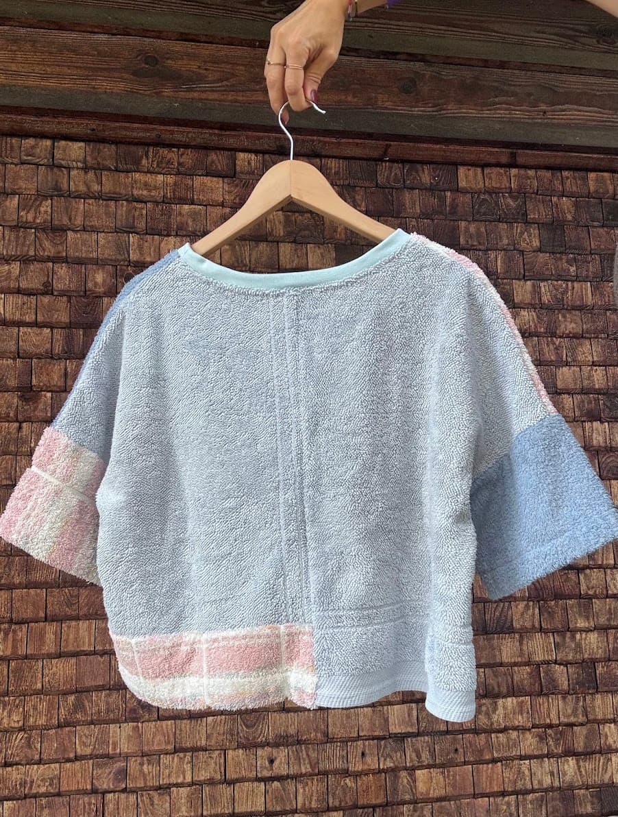 Rückansicht des Upcycling-Frotteeshirts „Blueberry '76“ von Old Wave an einem Kleiderbügel. Diese Perspektive betont die gespiegelte Materialaufteilung auf der Rückseite: Das Shirt ist größtenteils aus taubenblauem Frotteestoff gefertigt, während die linke Gesäßseite (vom Betrachter aus rechts) am unteren Saum einen dezenten Patch aus dem rosa-weiß karierten Vintage-Handtuchstoff aufweist.