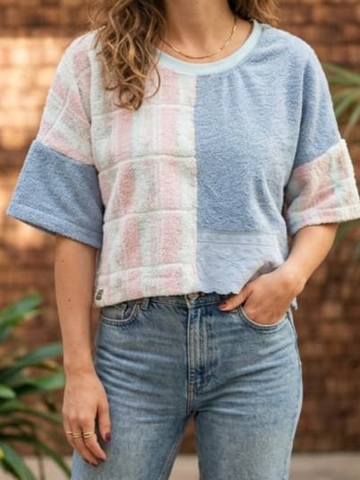 Detailansicht des Upcycling-Frotteeshirts „Blueberry '76“ von Old Wave an einem Model vor einem gemauerten Hintergrund. Das lockere Oversize-Oberteil zeigt ein asymmetrisches Patchwork-Design: Eine Hälfte besticht durch ein hellblau-rosa kariertes Handtuchmuster, während die andere Seite aus unifarbenem, hellblauem Vintage-Frotteestoff gefertigt ist. Dazu wird eine hellblaue Jeans getragen.