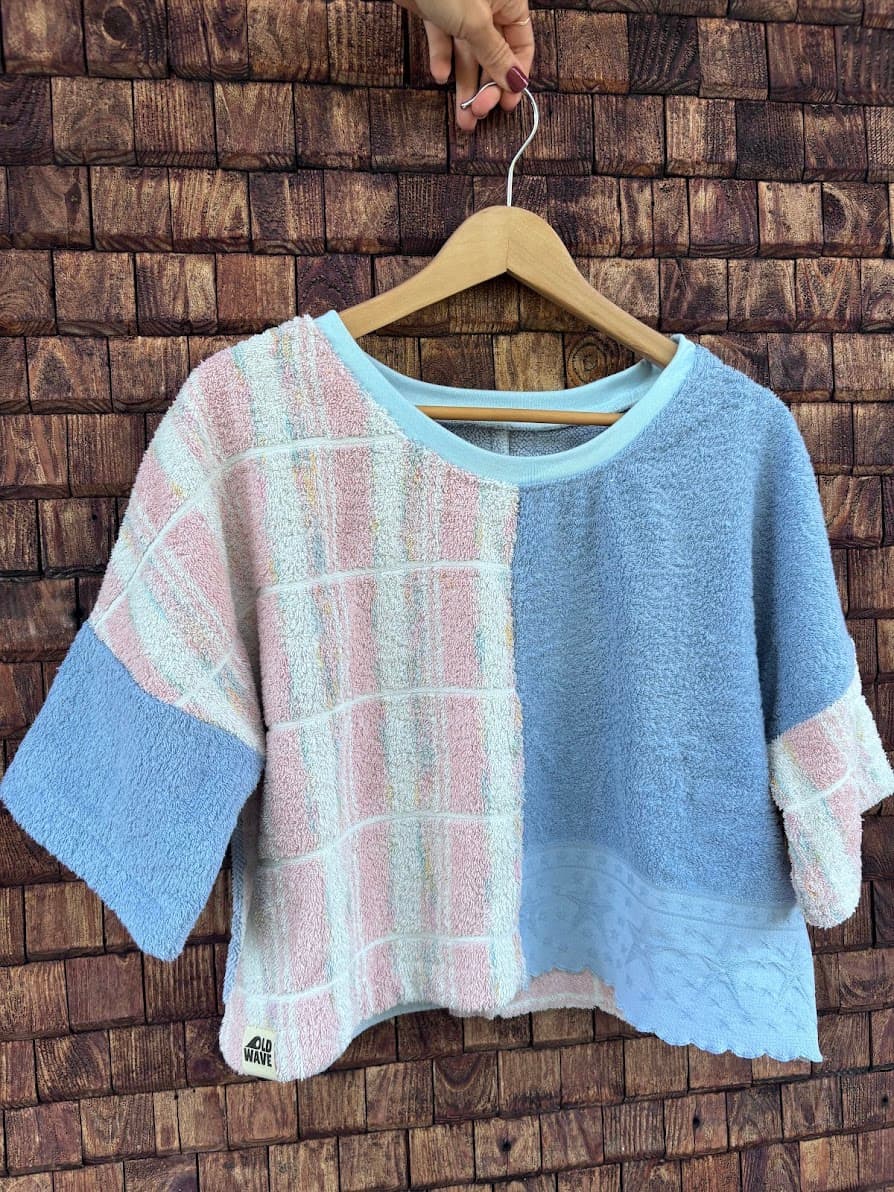 Vorderansicht des Towel Boxy Shirts „Blueberry '76“ von Old Wave auf einem Holzkleiderbügel vor einer dunklen Holzwand. Das kurzärmelige Upcycling-Oberteil im lockeren Boxy-Schnitt zeigt ein asymmetrisches Frottee-Patchwork-Design: Die linke Seite besteht aus einem weiß-rosa karierten Waffelstruktur-Handtuch, während die rechte Seite in schlichtem Taubenblau gehalten ist.