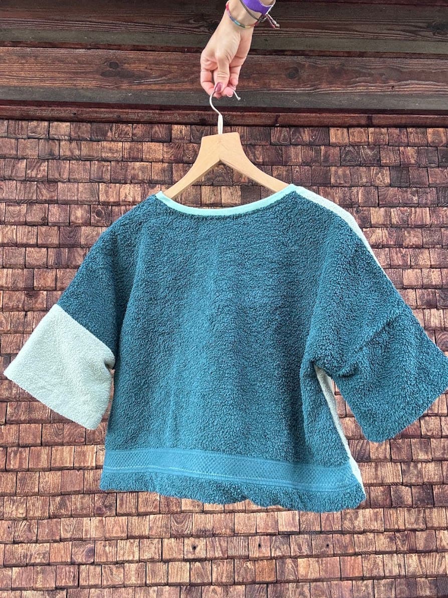 Rückansicht des Upcycling-Frotteeshirts „Coconut 73“ von Old Wave an einem Kleiderbügel. Diese Perspektive zeigt die Rückseite des weichen Handtuch-Oberteils, die fast vollständig aus dem tiefen, petrolblauen Frotteestoff besteht. Ein kontrastierender Akzent wird durch den linken Ärmel (vom Betrachter aus links) gesetzt, der aus einem hellbeigen Frotteestoff gefertigt ist.