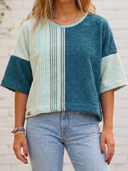 Nahaufnahme eines Models, das das Upcycling-Towel Boxy Shirt „Coconut 73“ von Old Wave trägt. Das kurzärmelige Oversize-Oberteil aus weichem Frotteestoff ist asymmetrisch gestaltet: Die linke Seite (vom Betrachter aus rechts) besteht aus tiefem Petrolblau, während die rechte Seite aus hellbeigem Handtuchstoff gefertigt ist und von einer vertikalen, mintgrün gestreiften Retro-Bordüre geziert wird. Dazu wird eine hellblaue Jeans getragen.