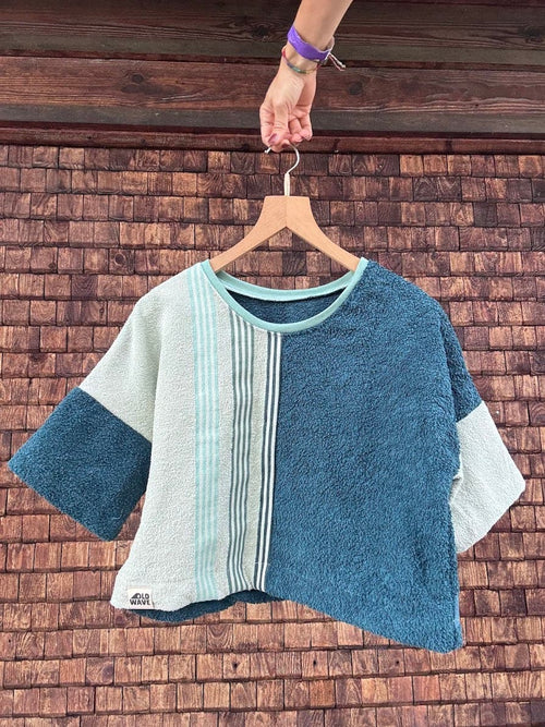 Vorderansicht des Towel Boxy Shirts „Coconut 73“ von Old Wave auf einem Holzkleiderbügel vor einer dunklen Holzwand. Das kurzärmelige Upcycling-Oberteil im lockeren Oversize-Schnitt besticht durch ein asymmetrisches Frottee-Design: Die rechte Vorderseite und der linke Ärmel sind aus einem petrolblauen Handtuchstoff gefertigt. Die linke Seite kombiniert ein hellbeiges Frottee-Element mit einer auffälligen, mintgrün gestreiften Vintage-Bordüre, die vertikal über die Brust verläuft.