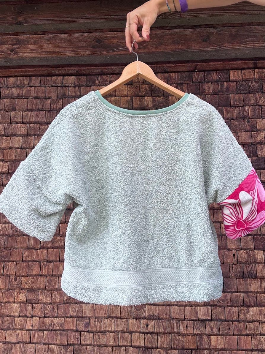 Rückansicht des Upcycling-Frotteeshirts „Lemon 65“ von Old Wave an einem Kleiderbügel. Diese Ansicht zeigt die schlichte, mintgrüne Rückseite des weichen Handtuch-Oberteils im lockeren Oversize-Schnitt, wobei am linken Ärmel das kontrastierende, intensiv pinkfarbene Blumenmuster der Vorderseite dekorativ hervorblitzt.