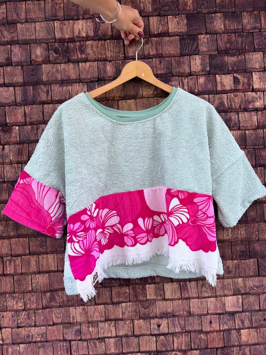 Vorderansicht des Towel Boxy Shirts „Lemon 65“ von Old Wave auf einem Holzkleiderbügel vor einer dunklen Holzwand. Das kurzärmelige Upcycling-Oberteil besteht aus mintgrünem Frotteestoff und wird am vorderen Saum durch eine breite, leuchtend pinkfarbene Stoffbahn mit weißen Südsee-Blumenmotiven und einem weißen Fransensaum akzentuiert.