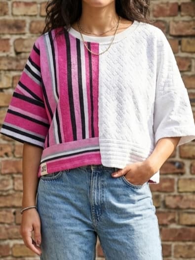 Detailansicht des Upcycling-Frotteeshirts „Strawberry '82“ von Old Wave an einem Model vor einer Backsteinwand. Das lockere Boxy Shirt zeigt ein asymmetrisches Design: Eine Hälfte besticht durch ein pink-schwarz gestreiftes Retro-Muster, die andere durch weißen Handtuchstoff mit Waffelstruktur. Dazu wird eine hellblaue Jeans getragen.