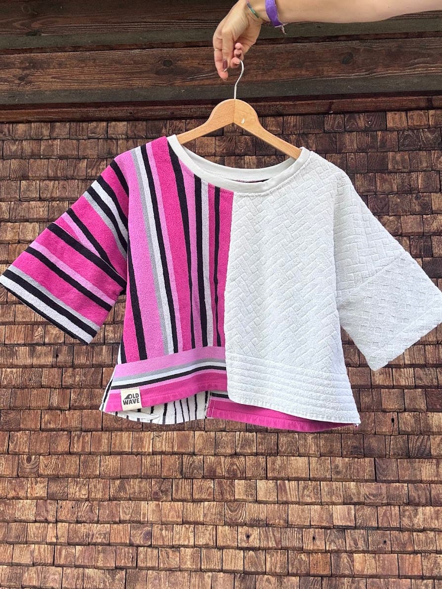 Vorderansicht des Towel Boxy Shirts „Strawberry '82“ von Old Wave auf einem Holzkleiderbügel vor einer dunklen Holzwand. Das kurzärmelige Upcycling-Oberteil im weiten Oversize-Schnitt teilt sich asymmetrisch auf: Die linke Seite (inklusive Ärmel) zeigt ein auffälliges Retro-Streifenmuster in Pink, Schwarz, Weiß und Grau, während die rechte Seite aus einem schlichten, weißen Handtuchstoff mit dezenter Waffelstruktur gefertigt ist.