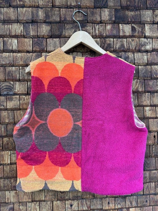 Rückansicht der gewendeten Frotteeweste 'Blueberry' von Old Wave. Zeigt das Patchwork-Design mit großen floralen Retro-Kreisen links und unifarbenem magentafarbener Frotteefläche rechts vor einer Holzwand.