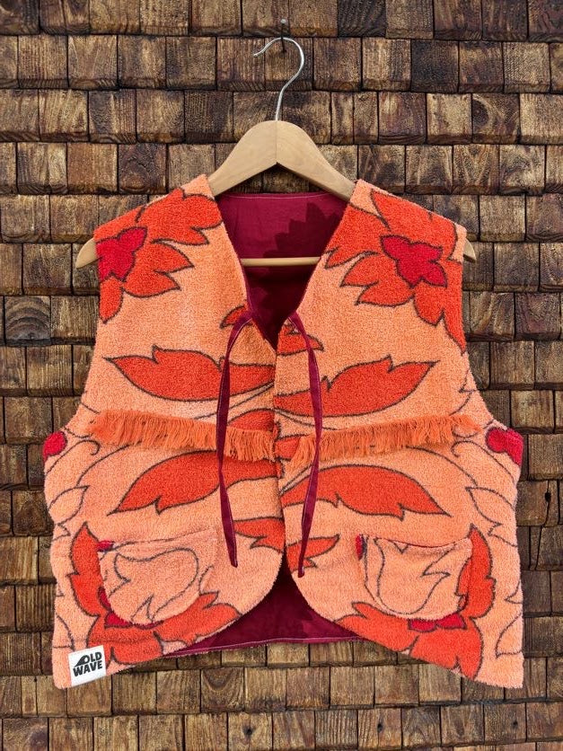 Vorderansicht der Frottee-Wendeweste 'Daisy' von Old Wave mit großflächigem Retro-Blumenmuster in Orange und Rot, Bindebändern und Old Wave Logo-Patch unten links vor einer Holzziegel-Wand.