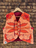Vorderansicht der Frottee-Wendeweste 'Daisy' von Old Wave mit großflächigem Retro-Blumenmuster in Orange und Rot, Bindebändern und Old Wave Logo-Patch unten links vor einer Holzziegel-Wand.