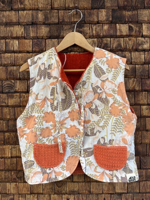 Vorderansicht der Frottee-Wendeweste 'Laguna' von Old Wave mit dichtem Vintage-Blumenmuster in Orange, Braun und Beige, zwei terrakottafarbenen Eingriffstaschen und Bindebändern vor einer Holzziegel-Wand.