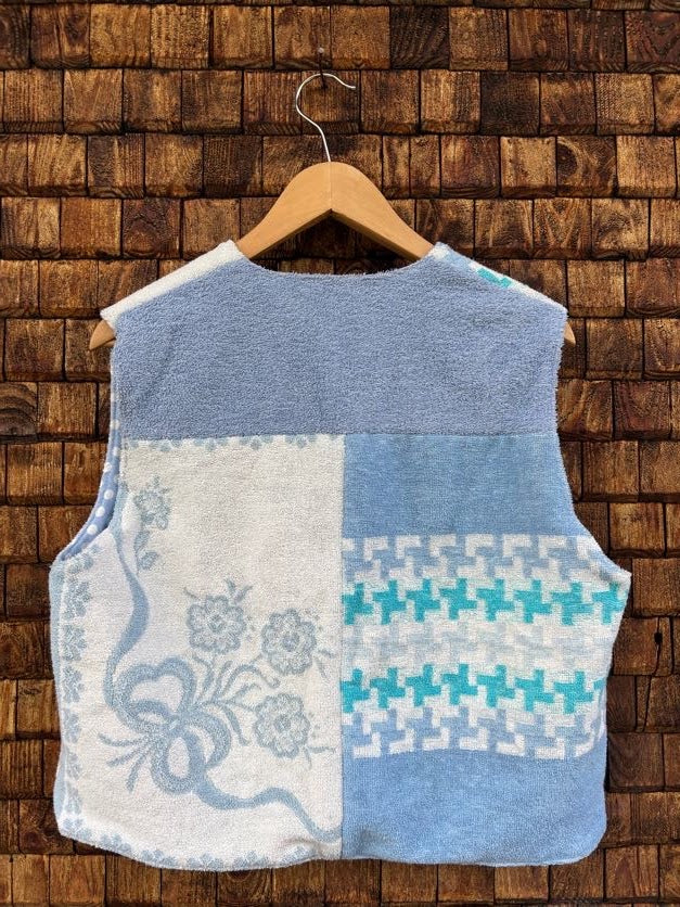 Rückansicht der gewendeten Frotteeweste 'Ocean' von Old Wave. Zeigt das viergeteilte Patchwork-Design aus unifarbenem Hellblau, weißem Frottee mit Blumenstickerei und Türkis-Hahnentritt-Muster vor einer dunklen Holzwand.