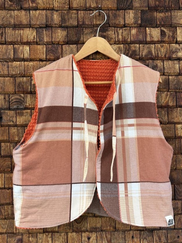 Vorderansicht der Frottee-Wendeweste 'Papaya' von Old Wave mit großflächigem Karomuster in Erdtönen, Braun, Terrakotta und Beige, Bindebändern und Logo-Patch unten rechts vor einer Holzziegel-Wand.