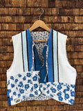 Vorderansicht der Frottee-Wendeweste 'Riviera' von Old Wave im maritimen Patchwork-Mix mit blauen Blumen-Prints, blau-weißen Streifen-Einsätzen und Bindebändern vor einer Holzziegel-Wand.
