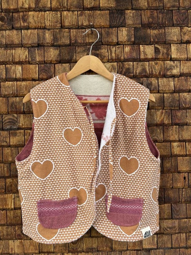 Vorderansicht der Frottee-Wendeweste 'Rose-Shell' von Old Wave mit hellbraun-weißen Feinstreifen, Herz-Motiven, zwei altrosafarbenen Eingriffstaschen und Logo-Patch unten rechts vor einer Holzziegel-Wand.