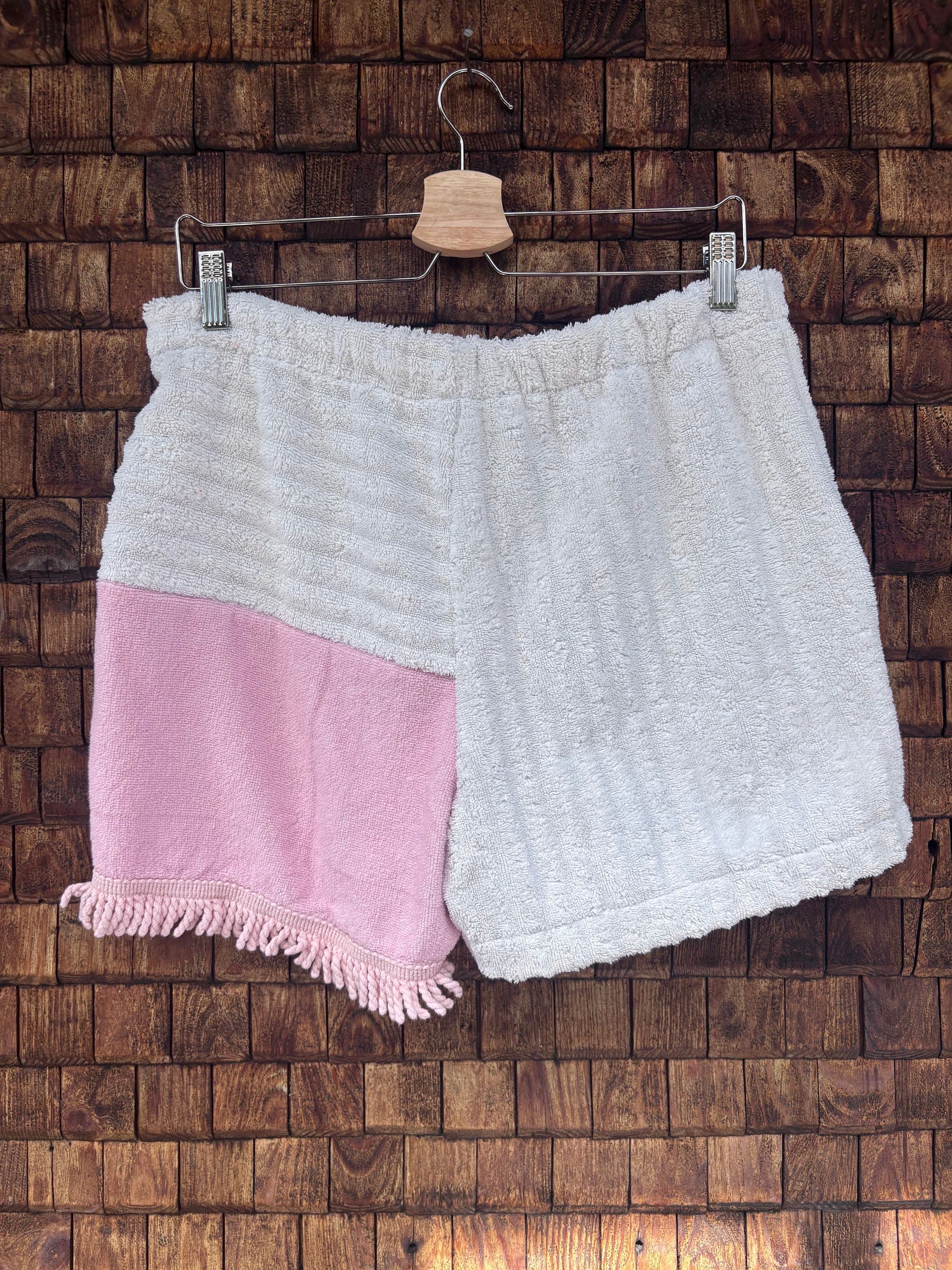 Rückansicht einer handgefertigten Pink salt '74 Shorts aus upgecycelten Vintage-Handtüchern in Weiß und softem Rosa mit Fransen-Saum, hängend vor einer braunen Holzwand.