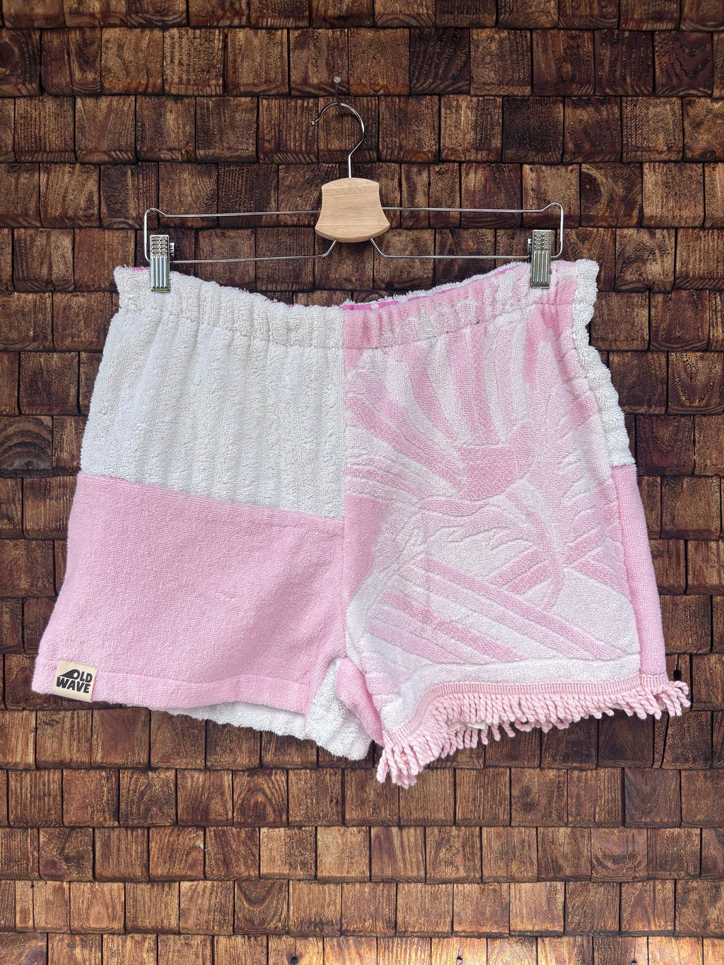 Vorderansicht der handgefertigten Pink salt '74 Shorts im Patchwork-Design aus Frottee-Stoff in Rosa und Weiß mit eingewebtem Vintage-Muster und Fransen-Details vor einer rustikalen Holzwand.