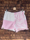 Vorderansicht der handgefertigten Pink salt '74 Shorts im Patchwork-Design aus Frottee-Stoff in Rosa und Weiß mit eingewebtem Vintage-Muster und Fransen-Details vor einer rustikalen Holzwand.