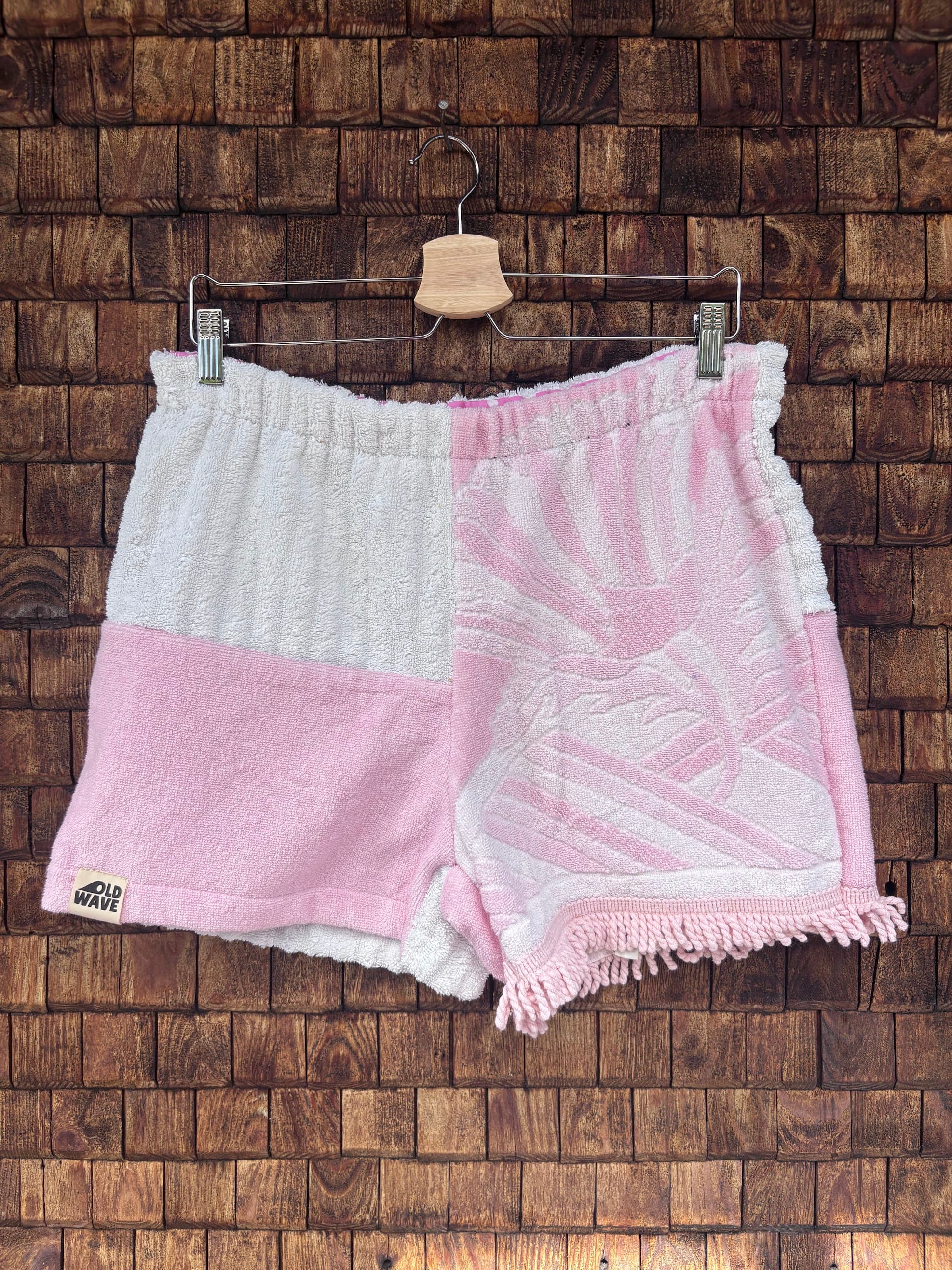 Vorderansicht der handgefertigten Pink salt '74 Shorts im Patchwork-Design aus Frottee-Stoff in Rosa und Weiß mit eingewebtem Vintage-Muster und Fransen-Details vor einer rustikalen Holzwand.