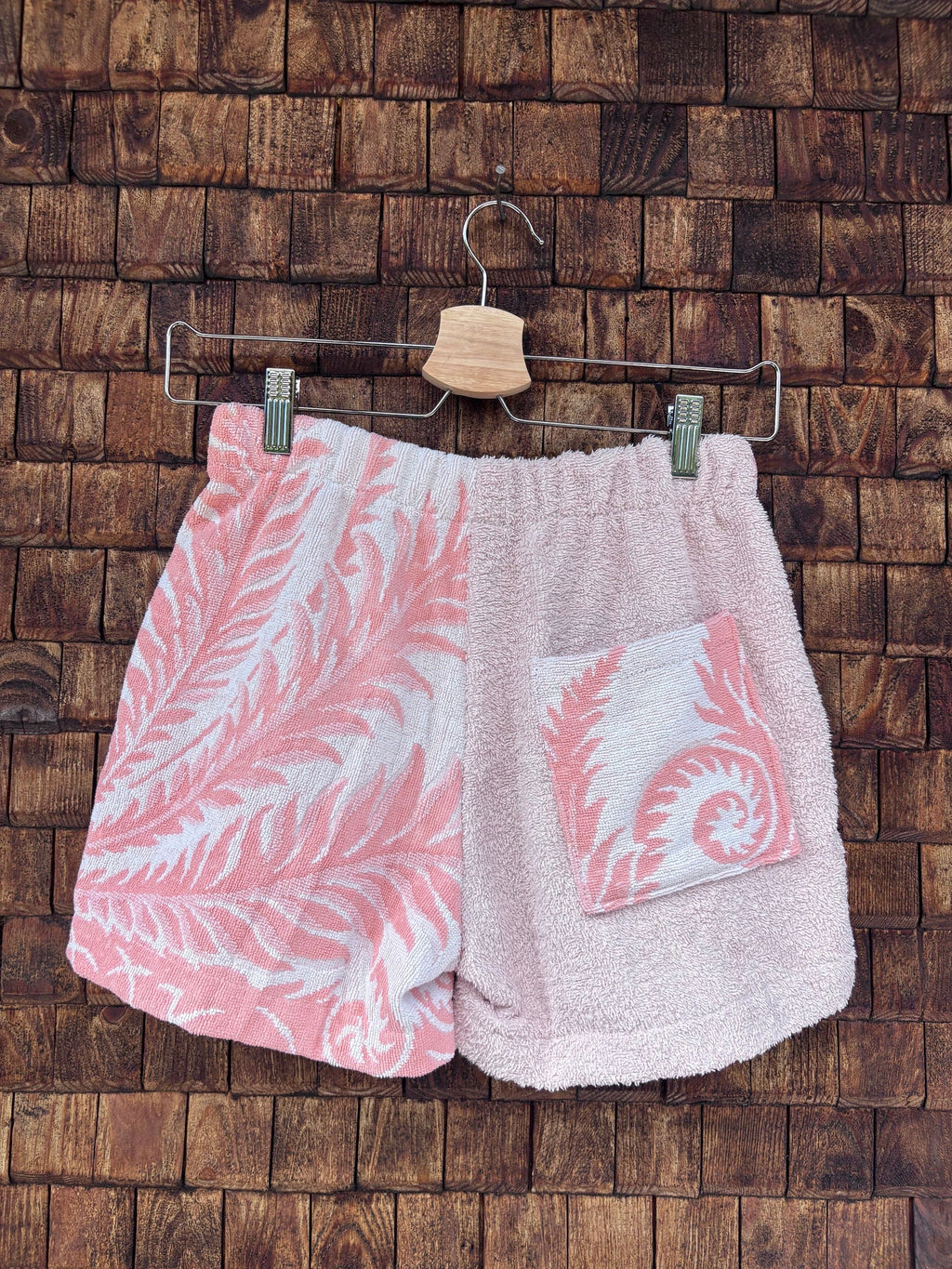 Rückansicht einer handgefertigten Riviera botanic '81 Shorts aus upgecycelten Vintage-Handtüchern. Linke Seite mit weiß-rosa Palmblatt-Muster, rechte Seite in zartem Hellrosa mit einer gemusterten, aufgesetzten Gesäßtasche, hängend vor einer Holzwand.