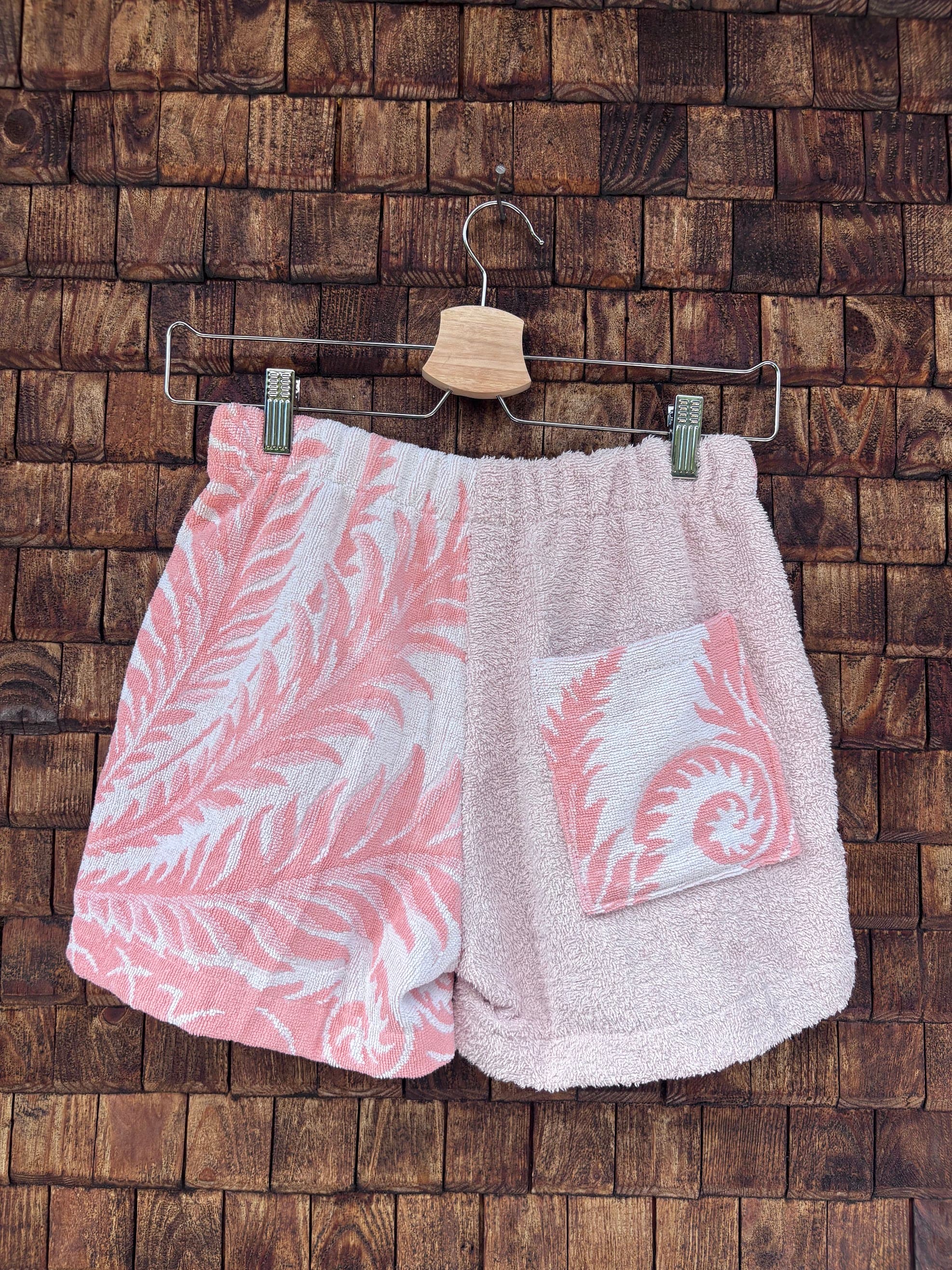 Rückansicht einer handgefertigten Riviera botanic '81 Shorts aus upgecycelten Vintage-Handtüchern. Linke Seite mit weiß-rosa Palmblatt-Muster, rechte Seite in zartem Hellrosa mit einer gemusterten, aufgesetzten Gesäßtasche, hängend vor einer Holzwand.