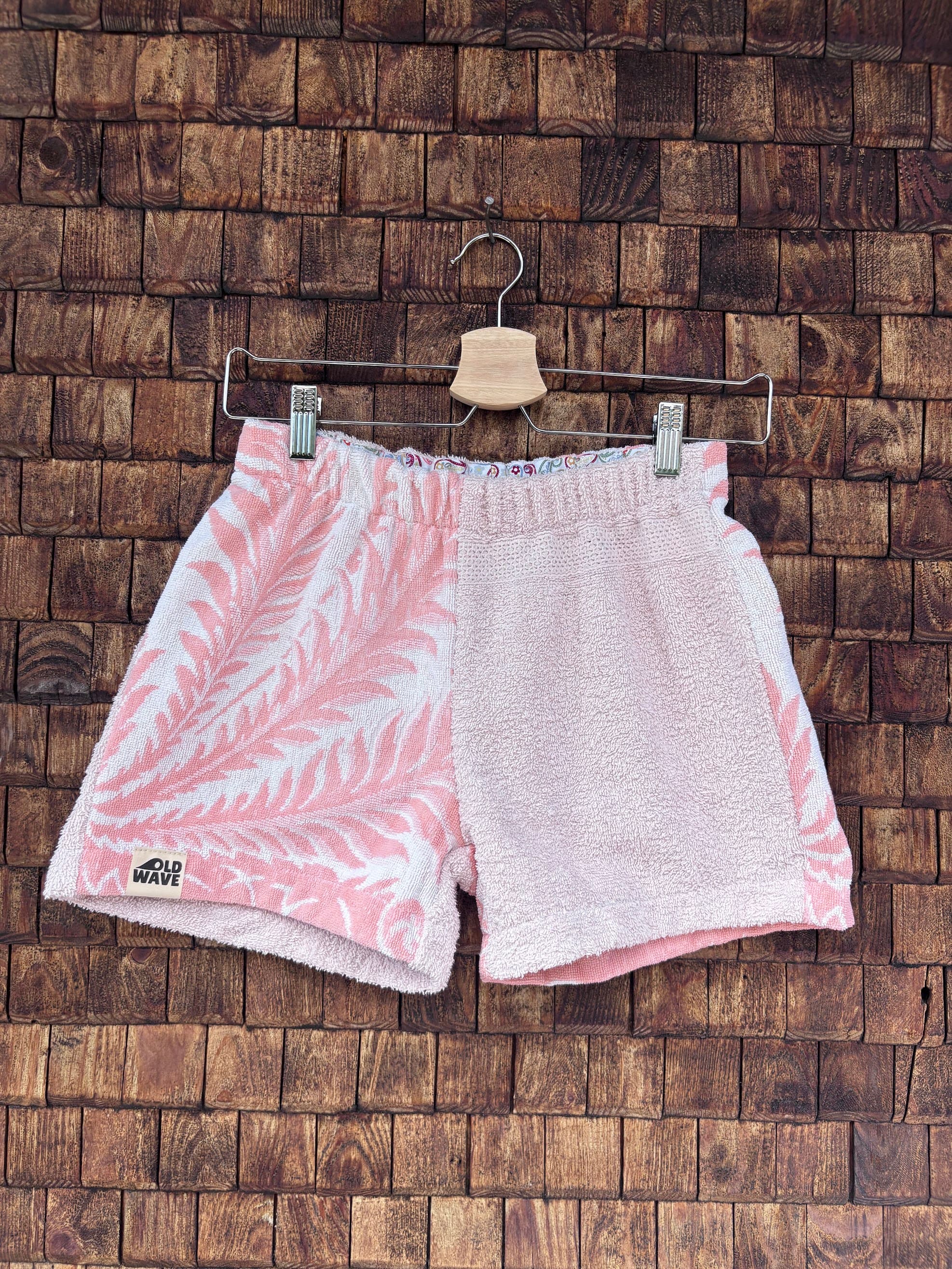 Vorderansicht der handgefertigten Riviera botanic '81 Shorts im Patchwork-Design aus Frottee-Stoff. Rechte Seite mit markantem botanischen Palmblatt-Muster in Rosa und Weiß, linke Seite in softem Pastellrosa vor einer rustikalen Holzwand.