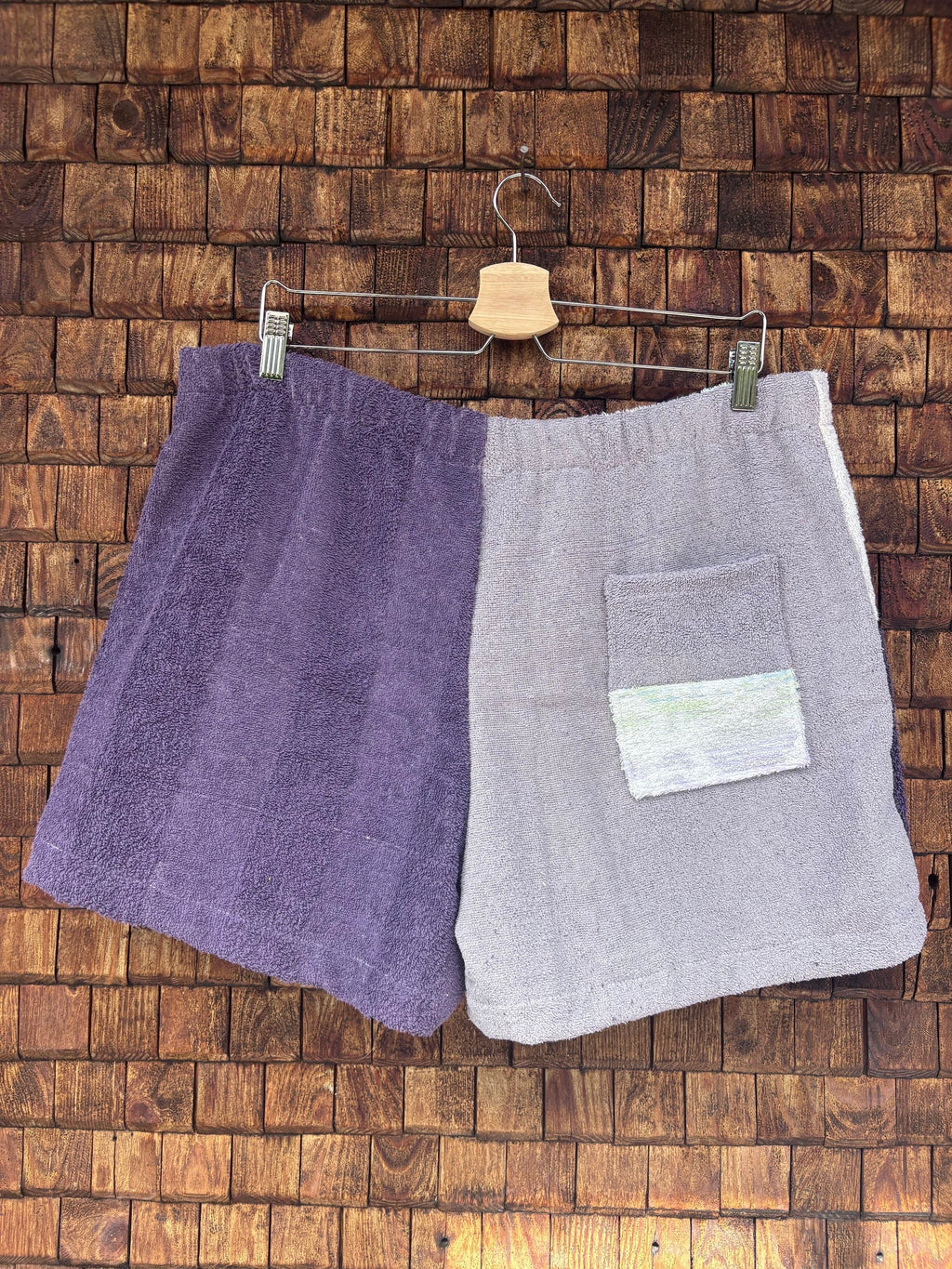 Rückansicht einer handgefertigten Riviera menthe '76 Shorts aus upgecycelten Vintage-Handtüchern in Flieder und Dunkellila mit einer aufgesetzten Gesäßtasche vor einer braunen Holzwand.