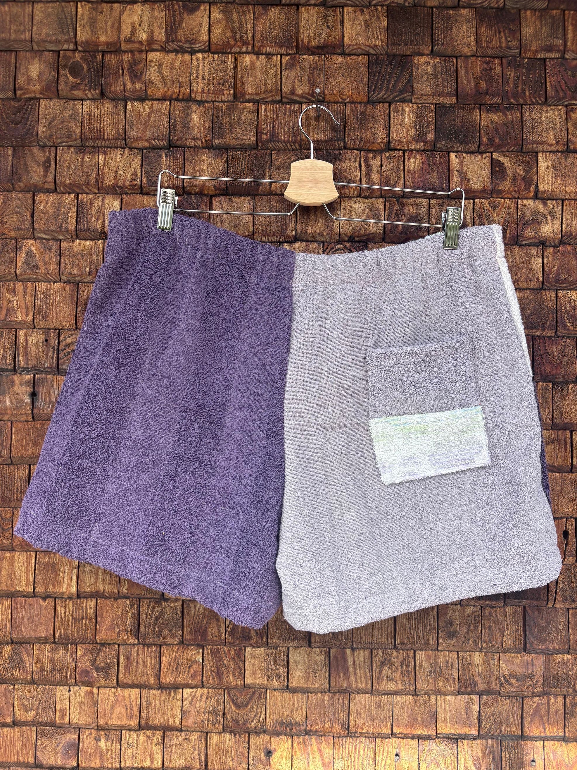 Rückansicht einer handgefertigten Riviera menthe '76 Shorts aus upgecycelten Vintage-Handtüchern in Flieder und Dunkellila mit einer aufgesetzten Gesäßtasche vor einer braunen Holzwand.