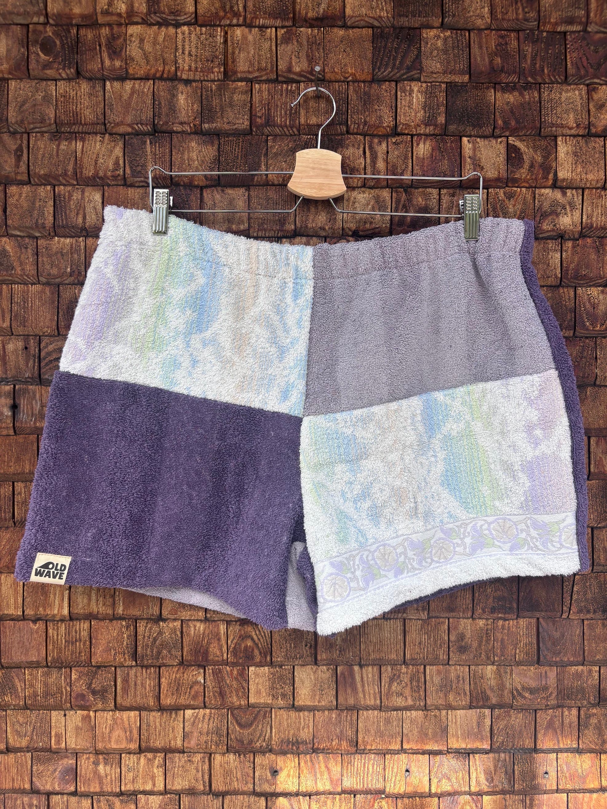 Vorderansicht der handgefertigten Riviera menthe '76 Shorts im Patchwork-Design aus Frottee-Stoff in Pastell-Lila und gemusterten Abschnitten, hängend vor einer rustikalen Holzwand.