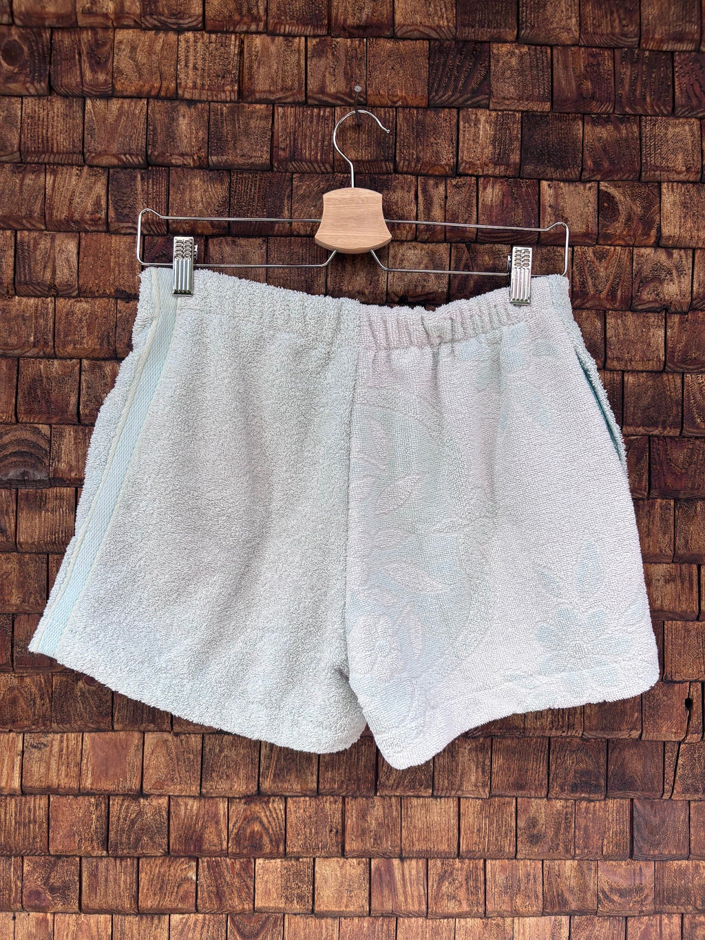 Rückansicht einer handgefertigten Riviera menthe '76 Shorts aus upgecycelten Vintage-Handtüchern in hellem Pastell-Mintgrün mit dezenter Frottee-Struktur, hängend vor einer braunen Holzwand.