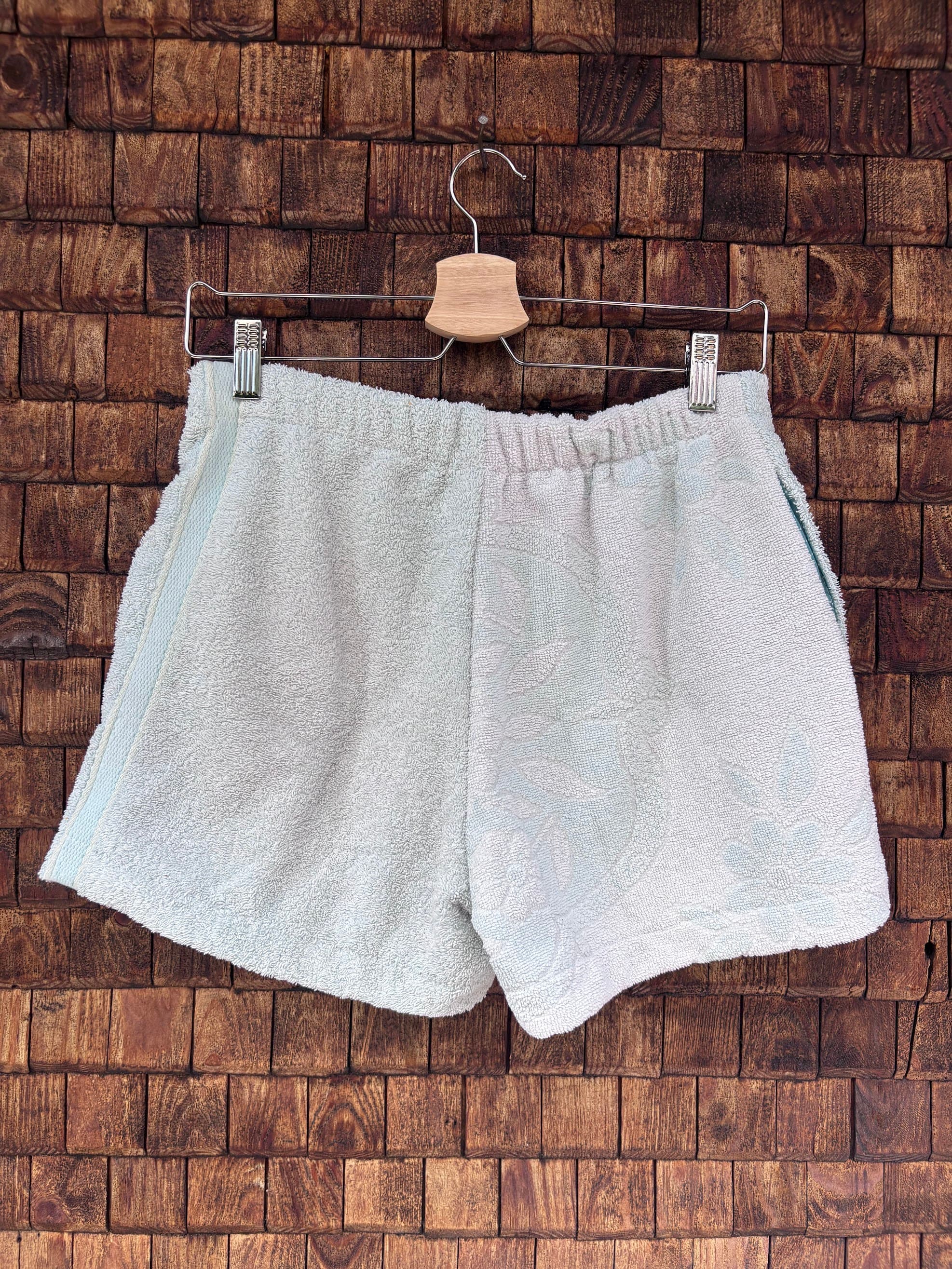 Rückansicht einer handgefertigten Riviera menthe '76 Shorts aus upgecycelten Vintage-Handtüchern in hellem Pastell-Mintgrün mit dezenter Frottee-Struktur, hängend vor einer braunen Holzwand.