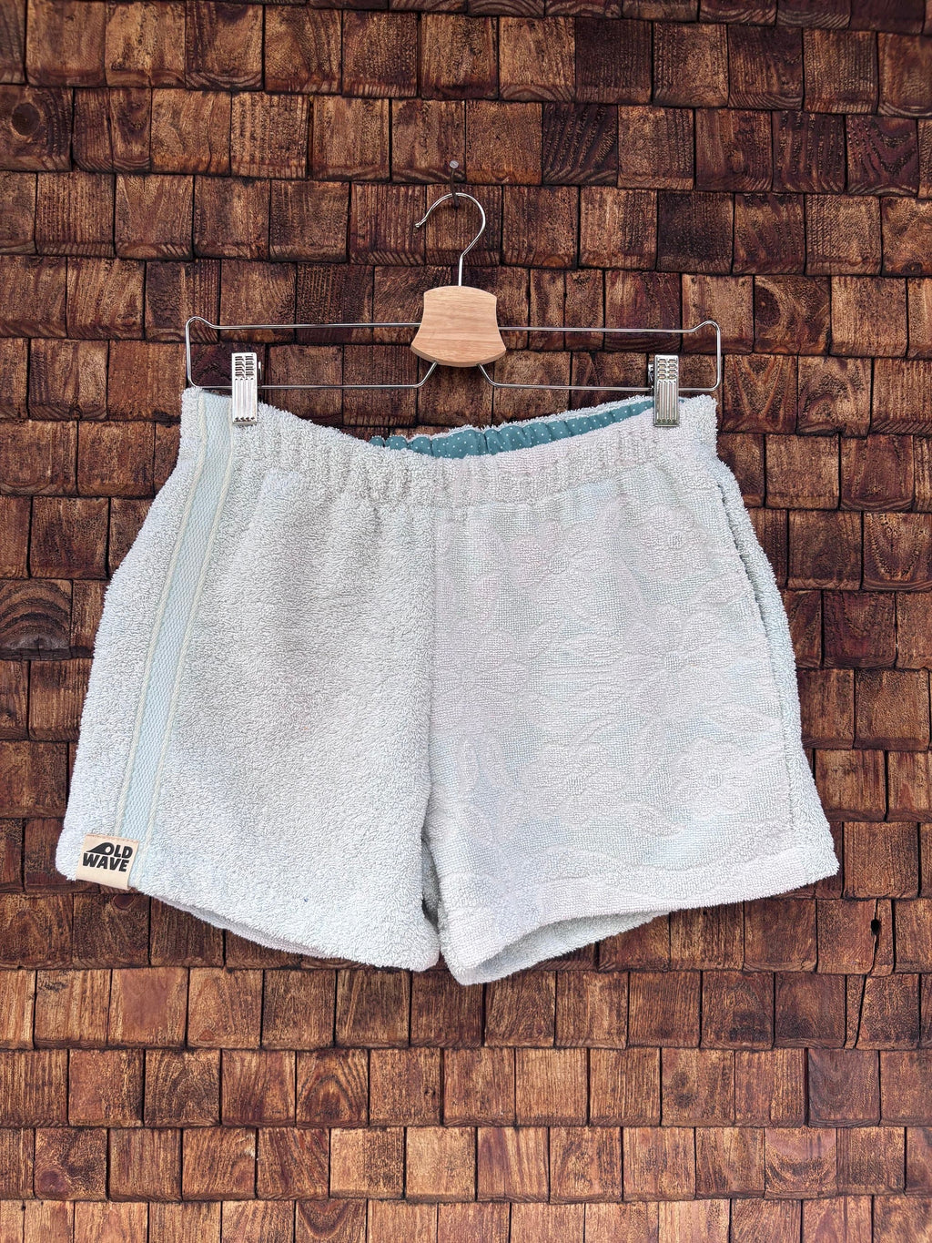 Vorderansicht der handgefertigten Riviera menthe '76 Shorts aus weichem Vintage-Frottee-Stoff in hellem Mintgrün, mit elastischem Bund und Logo-Patch am Saum, vor einer rustikalen Holzwand.