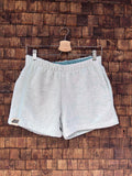 Vorderansicht der handgefertigten Riviera menthe '76 Shorts aus weichem Vintage-Frottee-Stoff in hellem Mintgrün, mit elastischem Bund und Logo-Patch am Saum, vor einer rustikalen Holzwand.