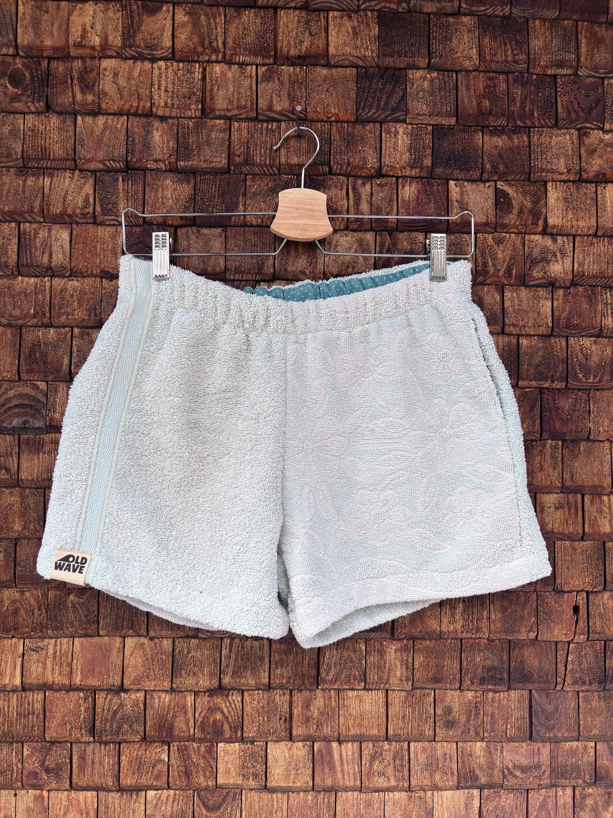 Vorderansicht der handgefertigten Riviera menthe '76 Shorts aus weichem Vintage-Frottee-Stoff in hellem Mintgrün, mit elastischem Bund und Logo-Patch am Saum, vor einer rustikalen Holzwand.