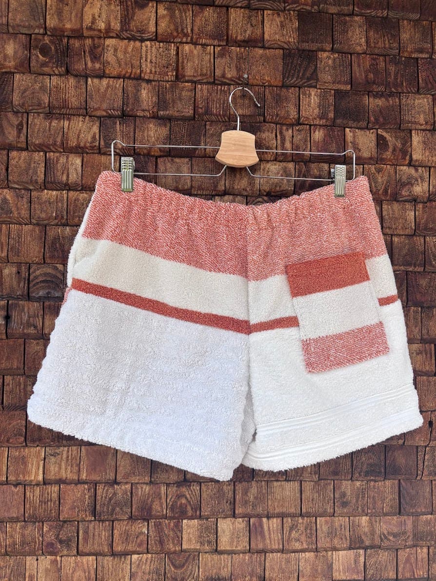 Rückansicht der handgefertigten Salty Coral ‘82 Upcycling-Shorts mit einem oberen Bundbereich in Korallenrot und einer markanten, aufgesetzten Gesäßtasche im koralle-weißen Streifendesign vor einer rustikalen Holzwand.
