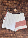 Vorderansicht der handgefertigten Salty Coral ‘82 Shorts aus upgecycelten Vintage-Handtüchern. Eine Beinhälfte zeigt strukturiertes weißes Waffel-Frottee, die andere Hälfte ist glatt-weiß mit einem breiten korallenroten Querstreifen, hängend vor einer Holzwand.