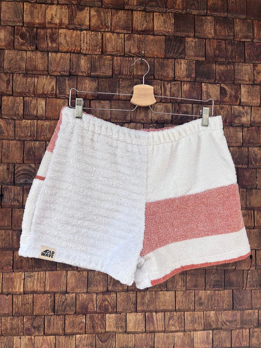 Vorderansicht der handgefertigten Salty Coral ‘82 Shorts aus upgecycelten Vintage-Handtüchern. Eine Beinhälfte zeigt strukturiertes weißes Waffel-Frottee, die andere Hälfte ist glatt-weiß mit einem breiten korallenroten Querstreifen, hängend vor einer Holzwand.