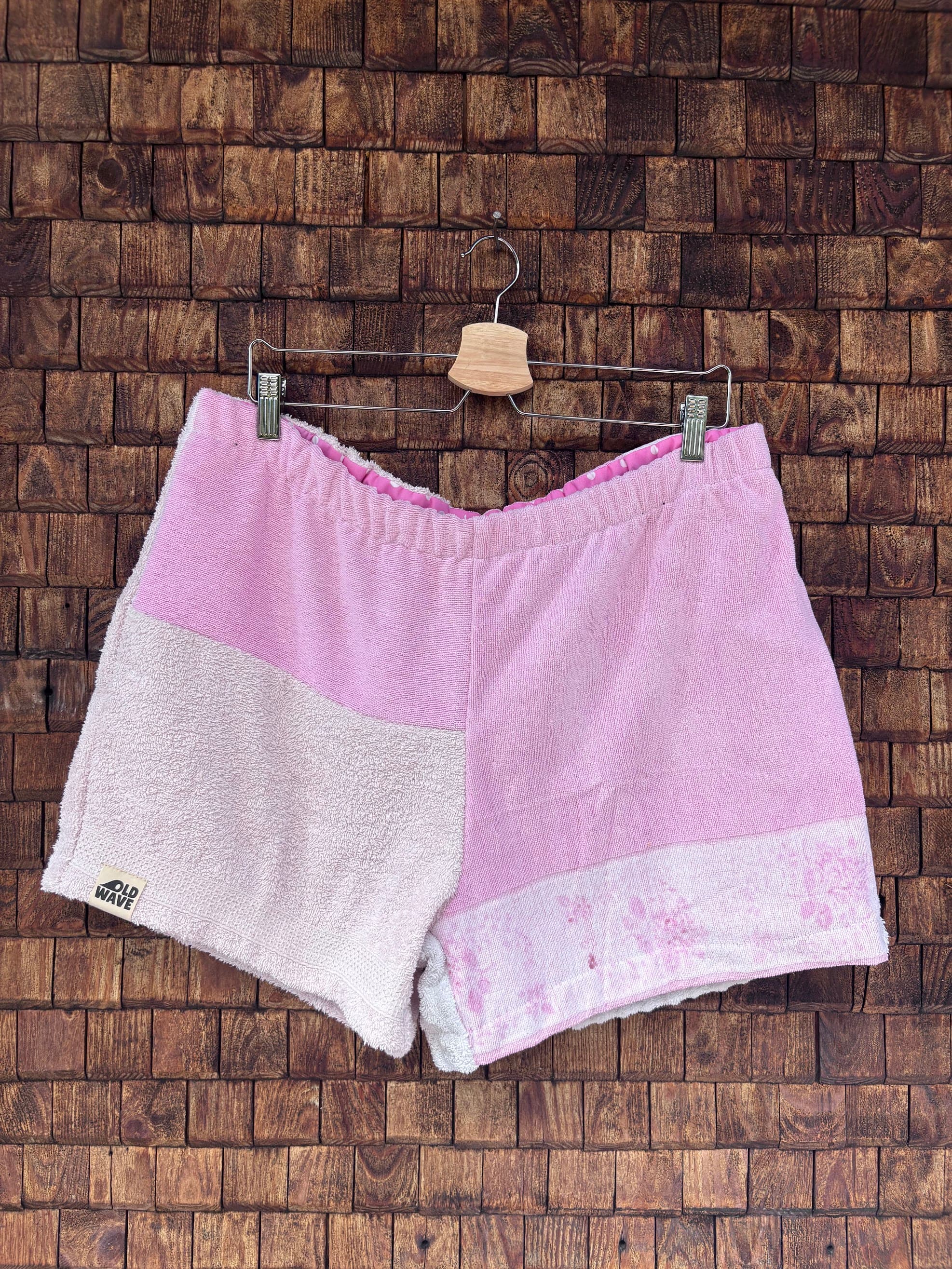 Vorderansicht der handgefertigten St. tropez pink '75 Shorts im Patchwork-Design aus Frottee-Stoff. Hauptteil in strahlendem Rosa, kombiniert mit einem helleren Pastellrosa und einem dezent gemusterten Saumabschnitt vor einer rustikalen Holzwand.
