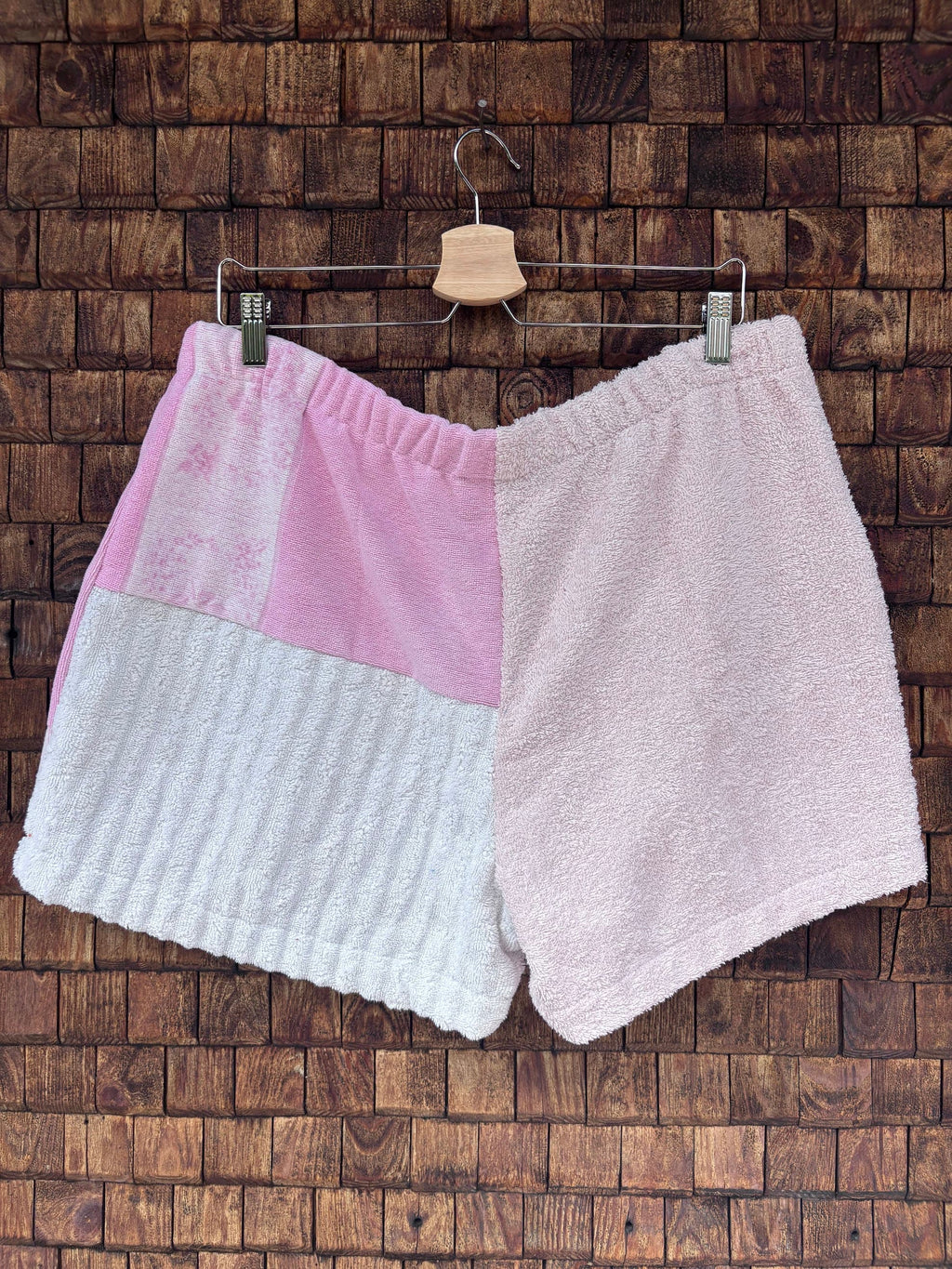 Rückansicht einer handgefertigten St. tropez pink '75 Shorts aus upgecycelten Vintage-Handtüchern. Rechte Seite in softem Pastellrosa, linke Seite unterteilt in leuchtendes Rosa und strukturiertes Weiß, hängend vor einer braunen Holzwand.