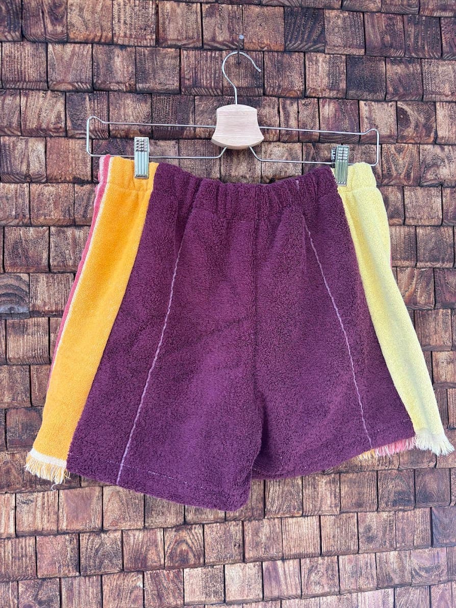 Rückansicht der handgefertigten Sunset Tile '83 Upcycling-Shorts, die das weinrote Frottee-Panel und die kontrastierenden gelben und blassgelben Seiten-Panels mit dem integrierten rosa Blumenmuster zeigen, hängend vor einer rustikalen Holzwand.