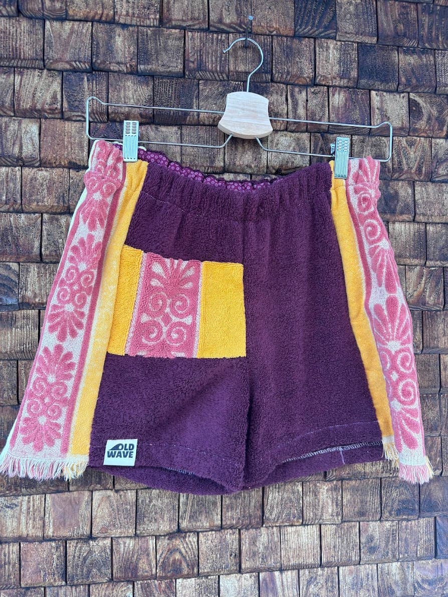 Vorderansicht der handgefertigten Sunset Tile '83 Shorts aus upgecycelten Vintage-Handtüchern. Zentrales Panel in Weinrot, kontrastierende gelbe Gesäßtasche mit rosa Blumenmuster und seitliche Panels mit auffälligem rosa Vintage-Blüten-Design, hängend vor einer Holzwand.
