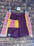 Vorderansicht der handgefertigten Sunset Tile '83 Shorts aus upgecycelten Vintage-Handtüchern. Zentrales Panel in Weinrot, kontrastierende gelbe Gesäßtasche mit rosa Blumenmuster und seitliche Panels mit auffälligem rosa Vintage-Blüten-Design, hängend vor einer Holzwand.