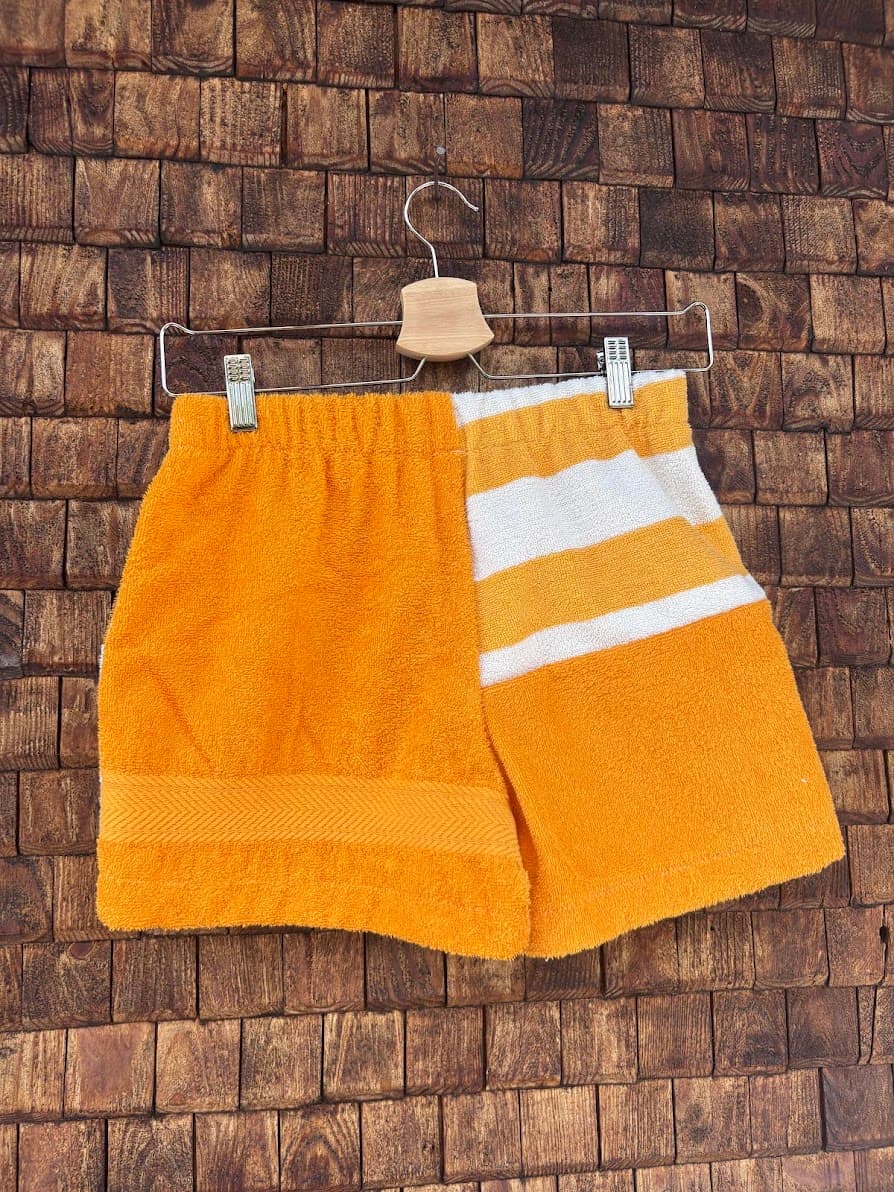 Rückansicht der Sunstripe ‘79 Upcycling-Shorts, hängend vor einer Holzwand. Eine Hälfte ist komplett in sattem Orange gehalten, während die andere Seite im oberen Bereich weiße Querstreifen zeigt.