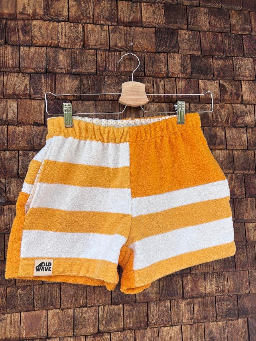 Vorderansicht der handgefertigten Sunstripe ‘79 Shorts aus upgecycelten Vintage-Handtüchern. Zu sehen ist das asymmetrische Design mit breiten orange-weißen Blockstreifen auf der einen und einem versetzten Streifenmuster auf der anderen Seite, hängend vor einer rustikalen Holzwand.