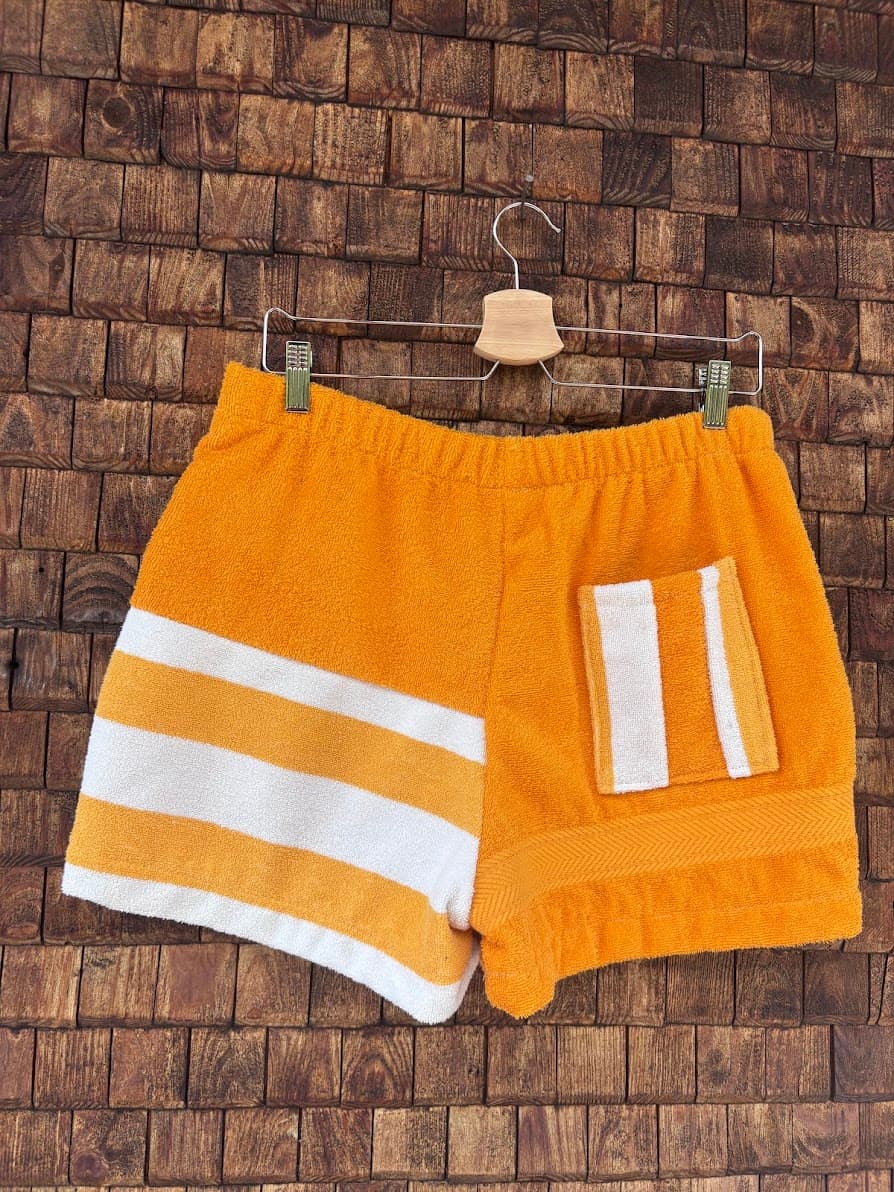 Rückansicht der handgefertigten Sunstripe Lounge ‘80 Upcycling-Shorts mit horizontal gestreifter linker Seite und einer kontrastierenden, vertikal gestreiften Gesäßtasche auf dem orangefarbenen Frottee-Panel vor einer rustikalen Holzwand.
