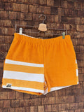 Vorderansicht der handgefertigten Sunstripe Lounge ‘80 Shorts aus upgecycelten Vintage-Handtüchern. Eine Seite in leuchtendem Orange, die andere Seite mit markanten, horizontalen Blockstreifen in Orange und Weiß, hängend vor einer Holzwand.