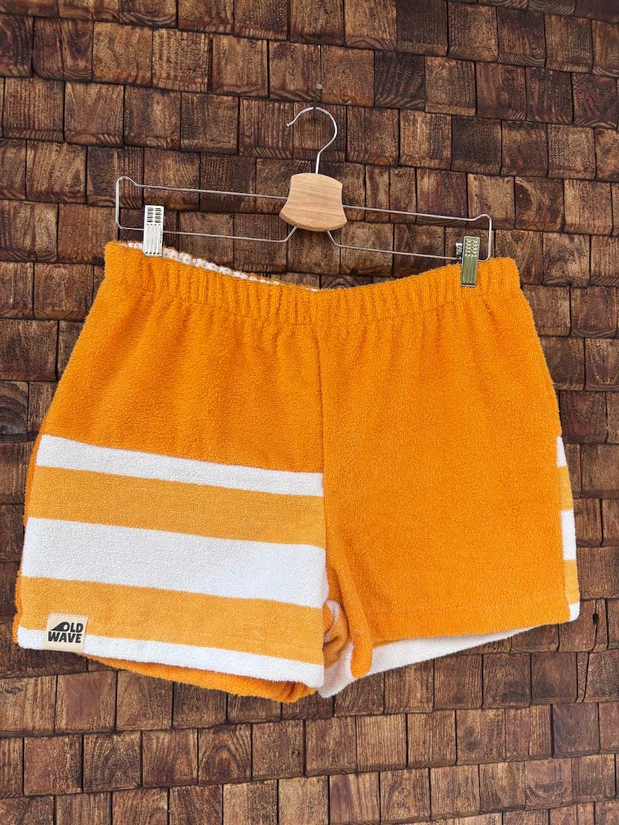 Vorderansicht der handgefertigten Sunstripe Lounge ‘80 Shorts aus upgecycelten Vintage-Handtüchern. Eine Seite in leuchtendem Orange, die andere Seite mit markanten, horizontalen Blockstreifen in Orange und Weiß, hängend vor einer Holzwand.